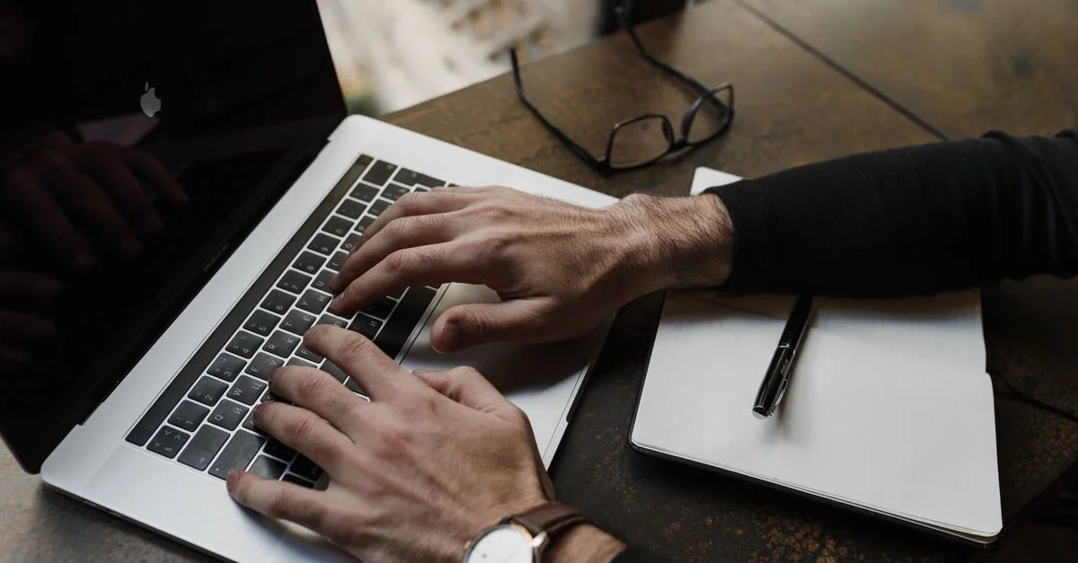 Vue concentrée des mains tapant sur un clavier d'ordinateur portable avec un bloc-notes et un stylo à proximité sur un bureau.